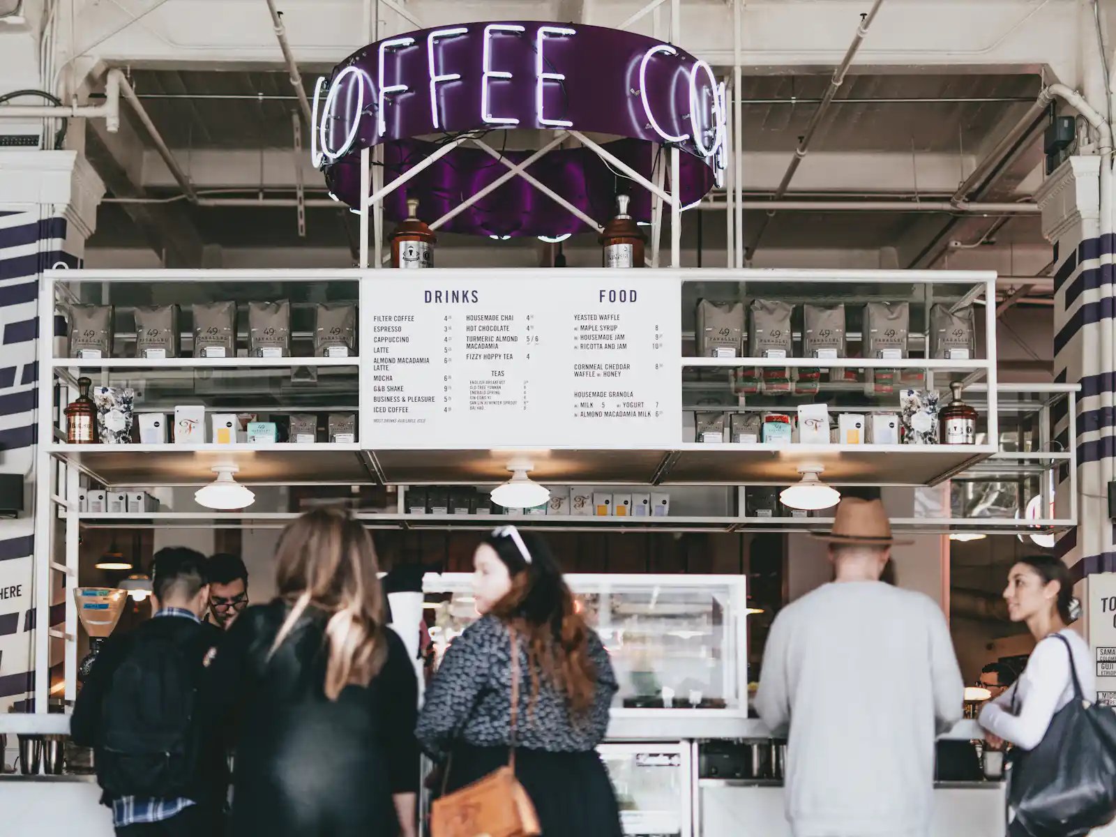 Ein belebtes Café mit einem modernen Design, einem beleuchteten "Coffee"-Schild und mehreren Kunden an der Theke. HANF SPAR!